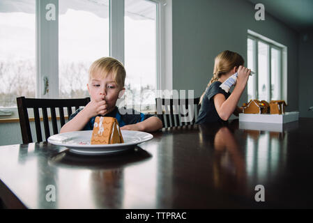 Frères et sœurs faisant maison en pain d'épices tout en étant assis à la maison pendant noël Banque D'Images