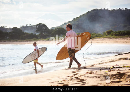 Vue latérale d'homme surfers carrying surfboards tandis que walking on beach Banque D'Images