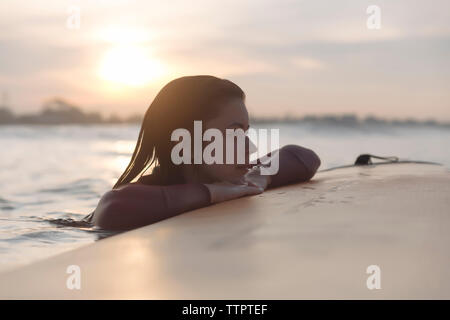 Young woman leaning on surf tout en regardant ailleurs en mer contre sky pendant le coucher du soleil Banque D'Images