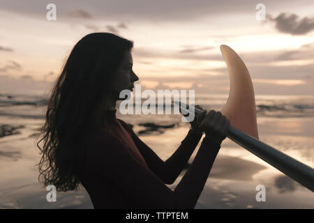 Vue latérale du young woman holding surfboard at beach debout contre le ciel au coucher du soleil Banque D'Images