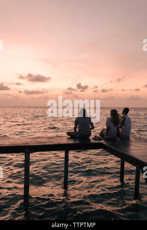 Silhouette friends sitting on pier sur la mer contre le ciel au coucher du soleil Banque D'Images