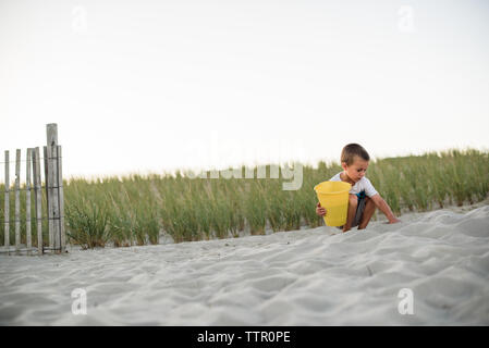 Jeune garçon de creuser dans le sable sur la plage avec un godet jaune au coucher du soleil Banque D'Images