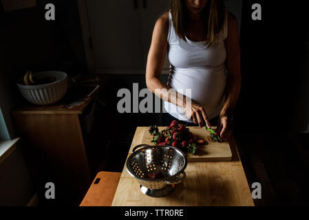 Future maman, couper les fraises dans la cuisine dans des conditions de faible luminosité Banque D'Images