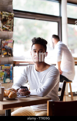 Portrait of young man with mobile phone ayant un croissant tout en étant assis au café Banque D'Images