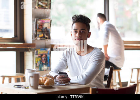 Portrait of young man holding mobile phone ayant des croissants et café au café Banque D'Images