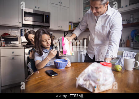 Père de verser les céréales petit déjeuner pour fille assise sur les genoux de sa mère dans la cuisine Banque D'Images