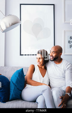 Smiling couple looking away while sitting on sofa contre le mur à la maison Banque D'Images
