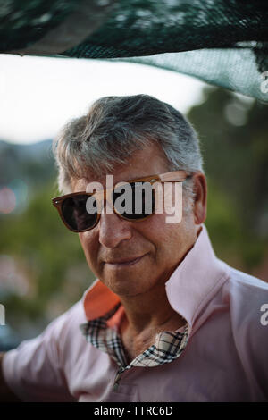 Close-up portrait of senior man wearing sunglasses en se tenant sur le terrasse de l'immeuble pendant le coucher du soleil Banque D'Images
