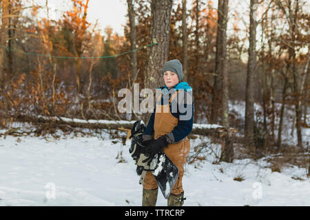Portrait of boy with snowboard debout sur le terrain couvert de neige dans la forêt Banque D'Images