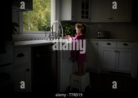 Side view of girl wearing cap rose se laver les mains dans un évier de cuisine en étant debout sur la selle à la maison Banque D'Images