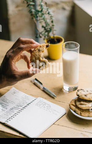 Cropped hand of man holding biscuit aux brisures de chocolat au lait et livre sur la table à la maison Banque D'Images