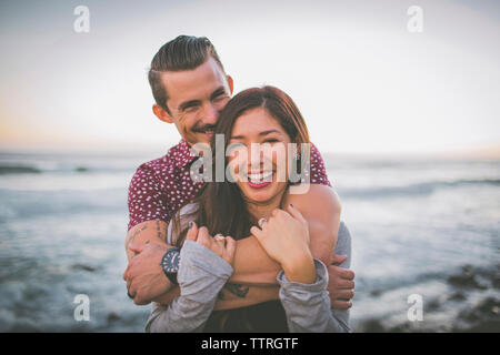 Portrait of happy couple standing at beach contre le ciel au coucher du soleil Banque D'Images