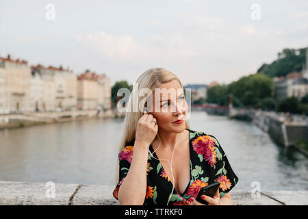 Femme à l'écoute de la musique tout en se tenant debout sur la passerelle sur la rivière contre ciel en ville Banque D'Images