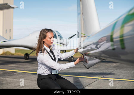 L'examen de pièces d'avion ingénieur femelle tandis que accroupi sur la piste de l'aéroport contre ciel lors de journée ensoleillée Banque D'Images