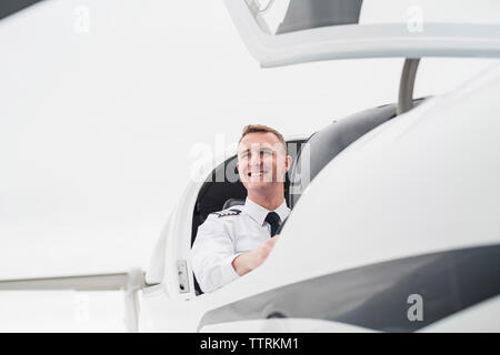 Low angle view of smiling male pilot in airplane assis contre le ciel à l'aéroport Banque D'Images