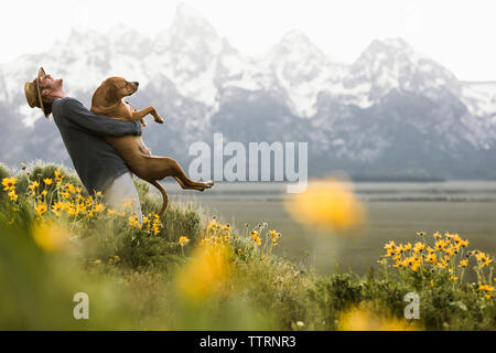 Vue latérale d'happy man carrying dog debout au milieu de plantes par contre le lac Montagne enneigée Banque D'Images