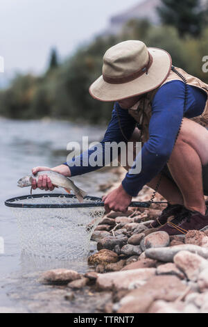 Vue de côté de l'homme tenant un poisson tout en accroupi sur des roches en rivière en forêt Banque D'Images