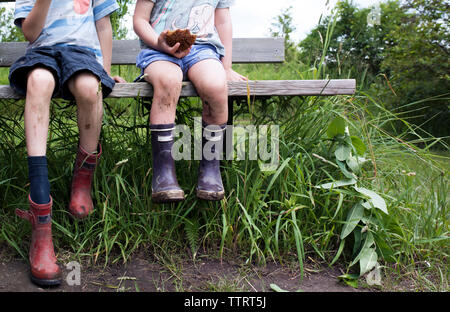 La section basse de la fratrie portant des bottes malpropres alors qu'assis sur un banc sur le terrain Banque D'Images