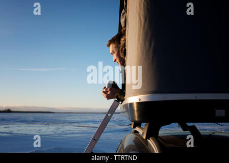 L'homme à l'écart tout en étant assis dans la tente de toit sur la voiture contre le ciel au coucher du soleil Banque D'Images