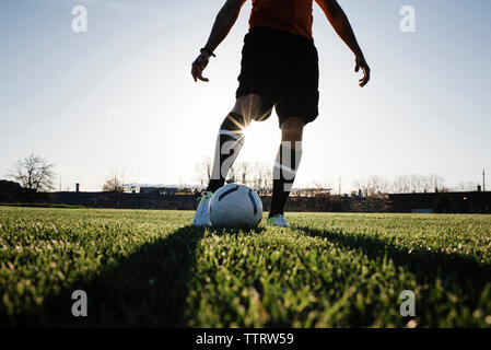 La section basse de l'homme jouant au football sur terrain herbeux contre ciel clair pendant le coucher du soleil Banque D'Images