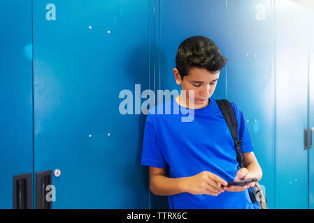 écolier utilisant un téléphone intelligent tout en se tenant contre un casier à l'école Banque D'Images