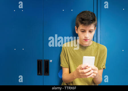 écolier utilisant un téléphone portable tout en se tenant debout contre un casier à l'école Banque D'Images