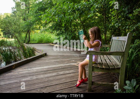 Vue latérale du girl reading book while sitting on bench at park Banque D'Images