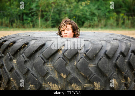 Portrait of cute boy peeking through dirty tire contre des plantes Banque D'Images