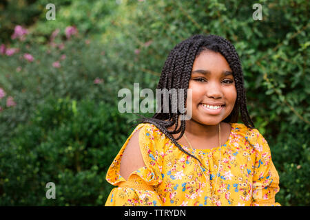 Portrait of smiling girl de tresses, assis contre des plantes dans le parc Banque D'Images