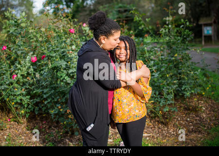 Mère embrassant sa fille sur le front en se tenant debout contre des plantes dans le parc