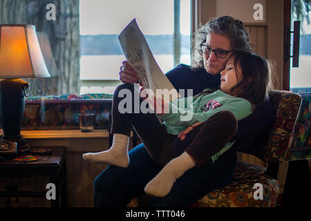 Une petite fille est assise sur les genoux de sa mère par une grande fenêtre Banque D'Images