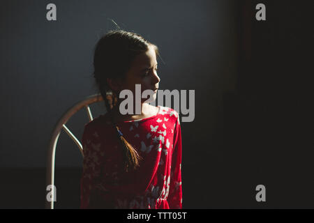 Une belle petite fille est assise sur une chaise dans la tache de lumière dans une pièce sombre Banque D'Images