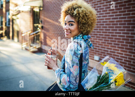 Portrait of smiling woman with ice tea et le bouquet walking on sidewalk Banque D'Images