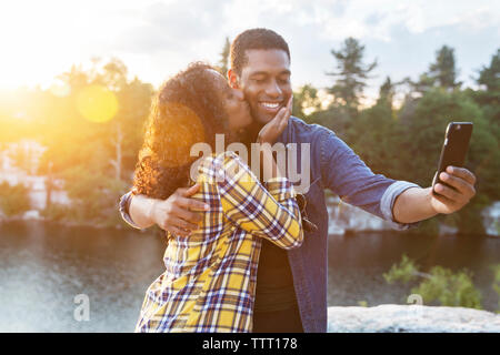 Woman kissing man prenant en selfies smart phone par le lac au coucher du soleil Banque D'Images