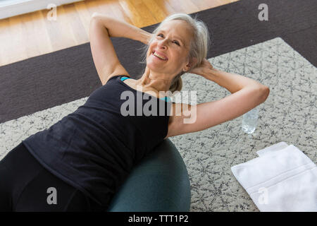 Smiling senior woman with hands behind head lying on fitness ball tout en exerçant à la maison Banque D'Images