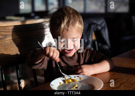 Close-up of cute happy boy de manger de la nourriture sur table en bois tout en restant assis à la maison Banque D'Images