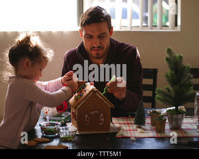 Fille avec le père decorating Gingerbread House sur la table à la maison Banque D'Images