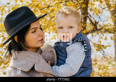 Mère portant mignon fils debout contre des arbres dans le parc en automne Banque D'Images