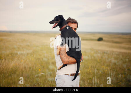 Portrait of man carrying dog en se tenant sur le terrain herbeux contre ciel nuageux Banque D'Images