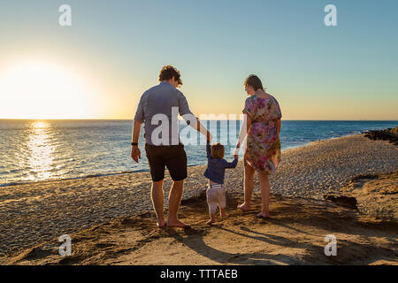 Vue arrière des parents tenant les mains de son fils alors qu'il marchait à la plage pendant le coucher du soleil Banque D'Images