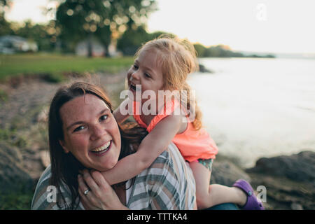 Portrait de mère heureuse étant embrassée par une fille mignonne tout en étant assis à la plage contre le ciel clair pendant le coucher du soleil Banque D'Images