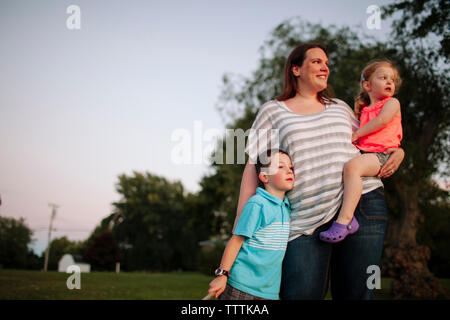 Mère souriante avec de jolis enfants qui regardent loin tout en se tenant contre le ciel dans le parc pendant le coucher du soleil Banque D'Images