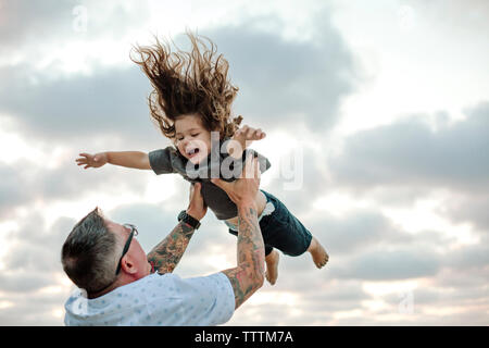 Low angle view of playful père fils jetant dans l'air tout en jouant contre ciel nuageux at beach Banque D'Images