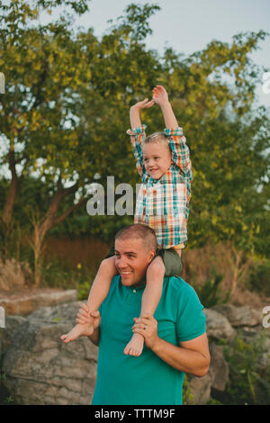 Happy father carrying son on shoulders debout contre des arbres pendant le coucher du soleil Banque D'Images