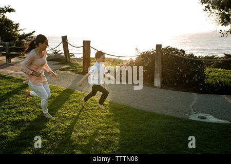 Mère courant derrière son on grassy field contre la mer au coucher du soleil Banque D'Images