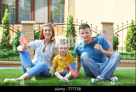 Famille heureuse avec peu de drapeaux américain assis sur l'herbe à côté de leur maison Banque D'Images