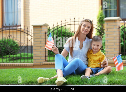 La mère et le fils avec peu de drapeaux américain assis sur l'herbe à côté de leur maison Banque D'Images