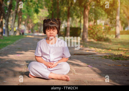 Les enfants asiatiques ou cute kid girl s'asseoir pour méditer en paix et vous détendre dans le pavillon de jardin à temple ou l'église et le port de robe blanche avec du soleil o Banque D'Images