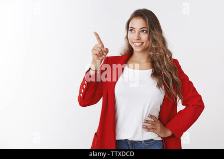 Studio shot magnifique aux cheveux bouclés 25s'woman wearing red veste élégante taille la main, pointant à heureux coin supérieur gauche smiling Banque D'Images