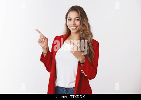 Charmant friendly-à female office worker aux cheveux bouclés, wearing red jacket, pointant l'index en haut à gauche smiling assertif, donnant Banque D'Images